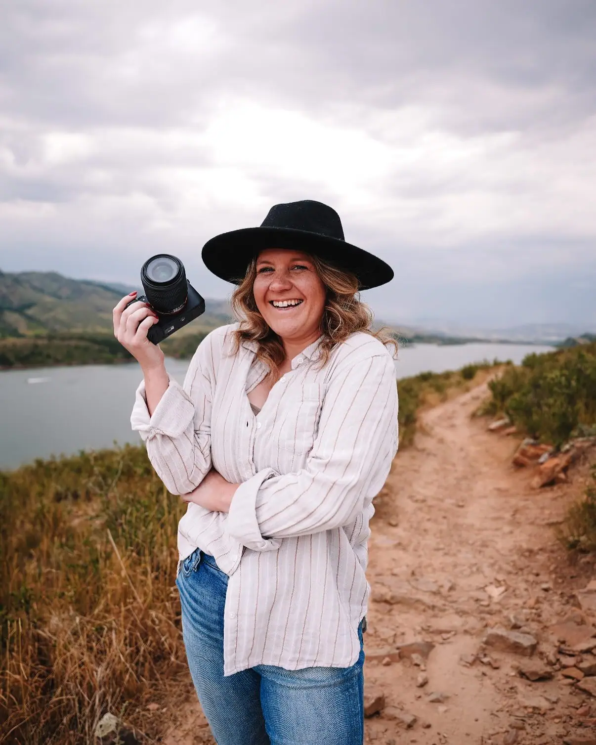 Delaney Ireland laughing on a hillside trail above a reservoir, camera raised in one hand