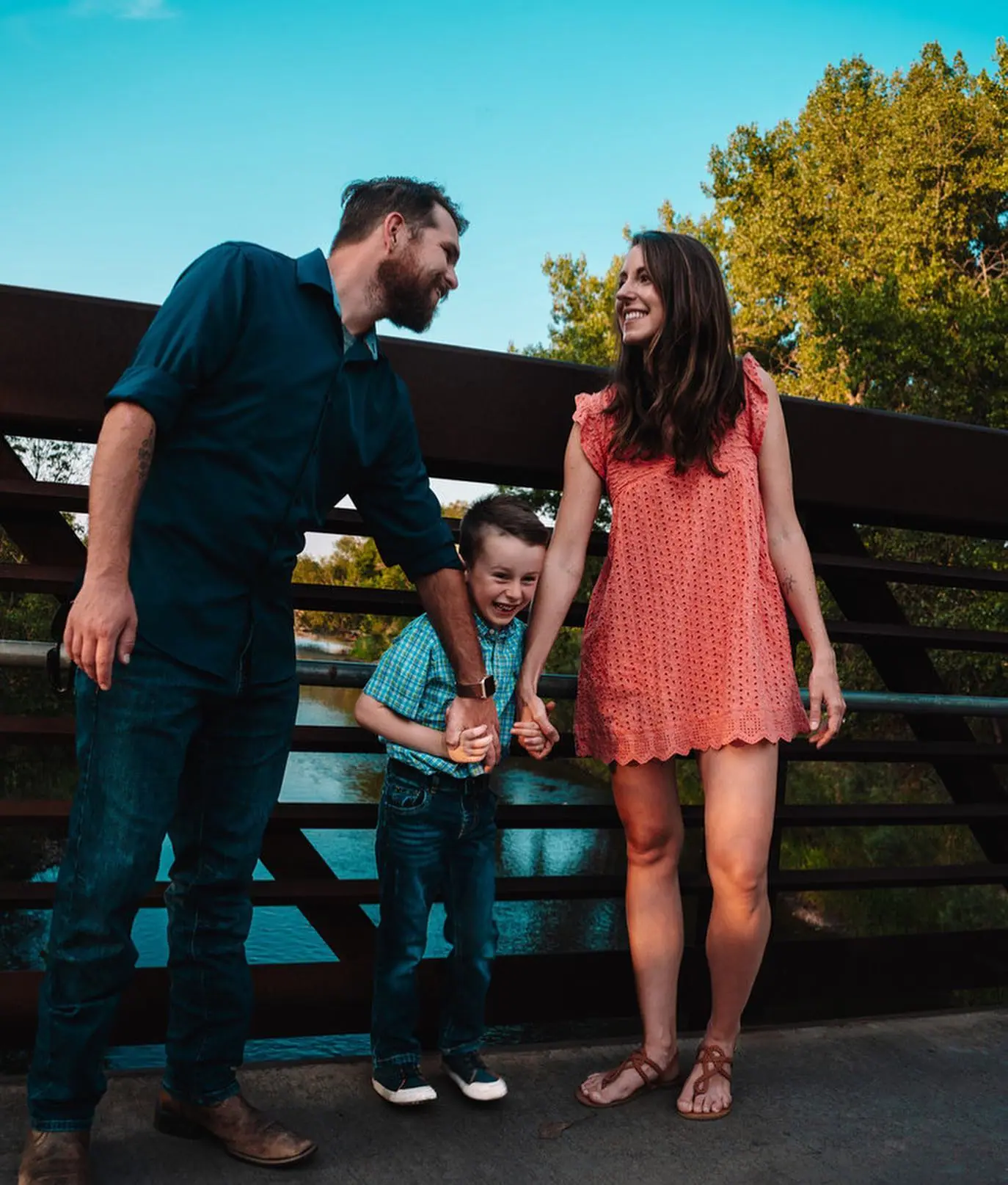 A family of three laughing together on a footbridge over a river at golden hour